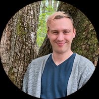 Rowan Nairn, a white person with short hair that's pink, blonde and brown, standing in front of a tree. They're wearing a dark blue v-neck t-shirt under a light-gray cardigan.